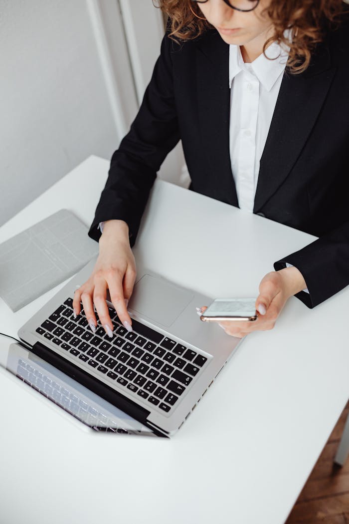 services-03 Focused businesswoman in black blazer using laptop and smartphone at desk.